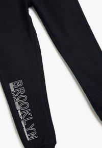 Czarne dresy z elastycznymi mankietami, z białym napisem „BROOKLYN” i gwiazdkami po boku. Miękki, teksturowany materiał dla wygody.