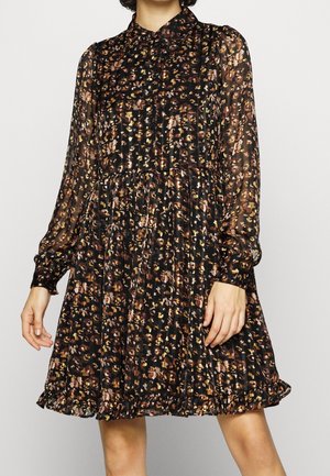Kvinde iført en sort langærmet kjole med krave, brune og beige blomsterprint og flæsekant.