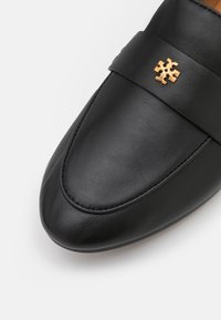 Mocassin en cuir noir avec une texture lisse, un bout rond et un accent en émail doré sur le devant. Présente un détail cousu le long de la semelle.