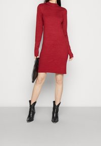 Rode gebreide jurk met lange mouwen en een hoge hals, met ribbelmanchetten en een getailleerde pasvorm, gecombineerd met een zwarte clutch en enkellaarsjes.