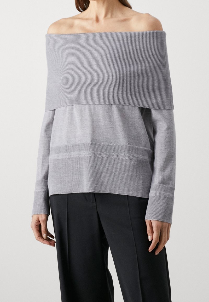Femme portant un pull en maille côtelée gris clair à épaules dénudées, associé à un pantalon noir à jambes larges sur fond uni.
