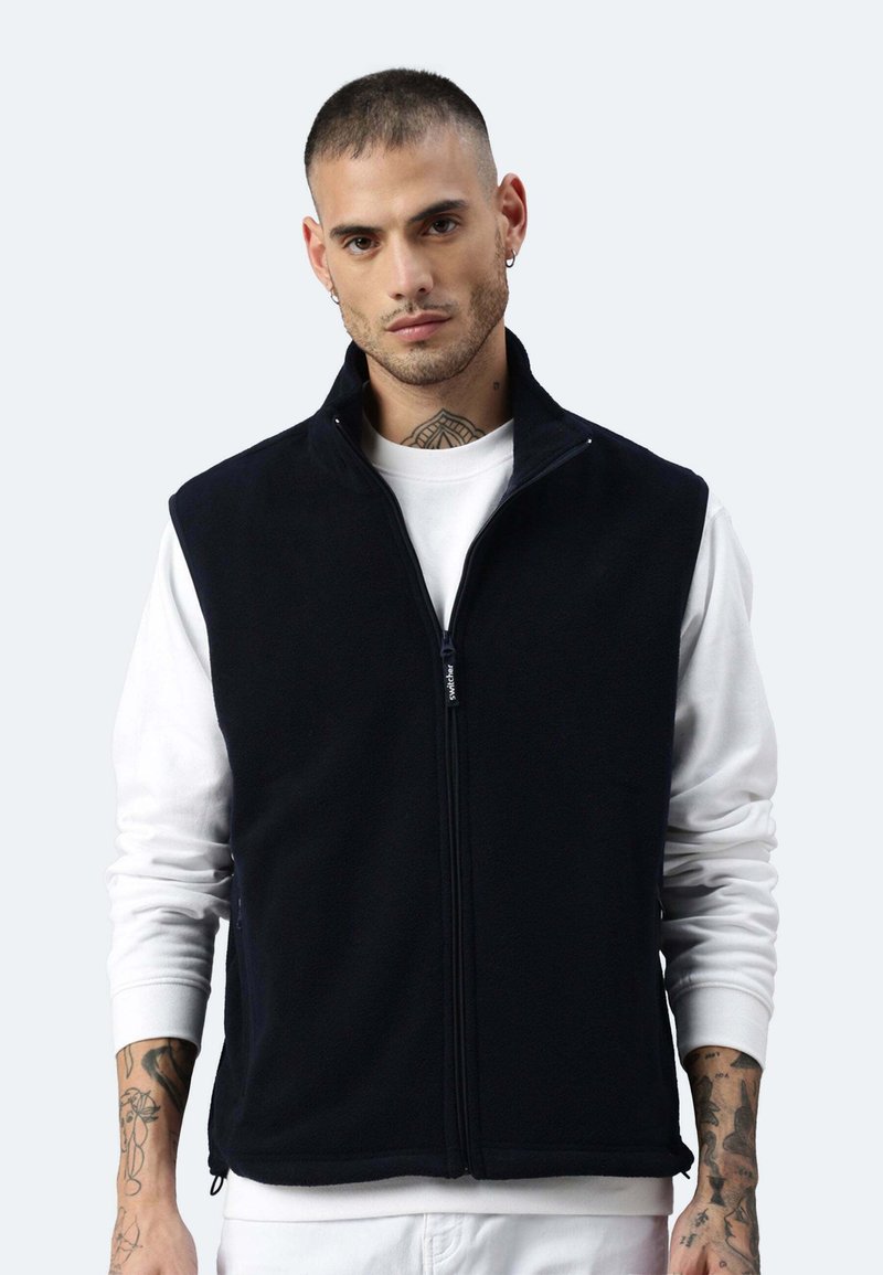 Schwarze Fleece-Weste mit hohem Kragen und durchgehendem Reißverschluss, getragen über einem weißen Langarmshirt. Verfügt über seitliche Taschen und eine lockere Passform.