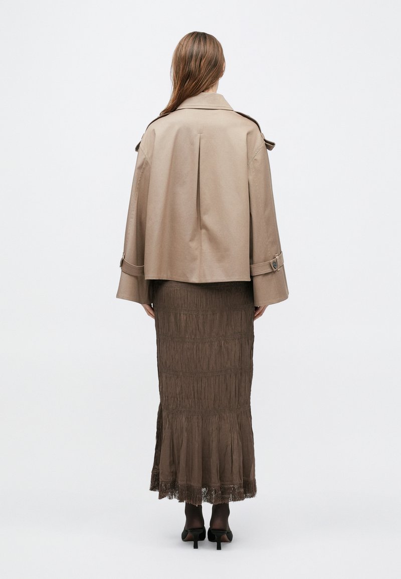Trench-coat beige à coupe ample, manches larges et détails de ceinture, associé à une jupe maxi marron texturée avec un ourlet à franges.
