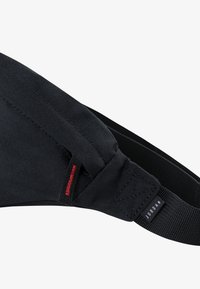 Correia de ombro em tecido preto com textura geométrica, apresentando detalhes em vermelho e branco e uma etiqueta de logo visível. Projetada para conforto e durabilidade.