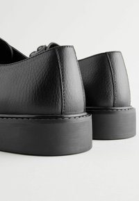 Chaussures en cuir noir avec une finition texturée, dotées d'une semelle épaisse noire et de détails de couture soignés. Accent sur le talon et la vue latérale.