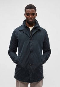 Casaco azul-marinho de comprimento médio, feito de um tecido suave. Apresenta colarinho, fecho de botão e bolsos laterais. Design simples sem padrões visíveis.