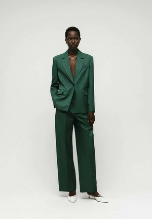 Groene op maat gemaakte pak met een blazer met pieklapels, brede broekspijpen en witte puntige hakken. Gladde textuur zonder zichtbare patronen.