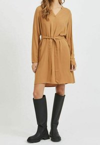 Robe marron à manches longues avec un col en V et une ceinture à la taille, associée à des bottes en caoutchouc noires montantes jusqu'au genou. Texture lisse, coupe décontractée, longueur mi-longue.