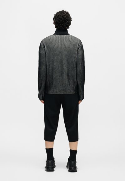 Άτομο με σγουρά μαλλιά στέκεται με την πλάτη, φοράει μαύρο και γκρι ριγωτό πουλόβερ, μαύρο παντελόνι μέχρι τον αστράγαλο, μαύρες κάλτσες και μαύρα παπούτσια.