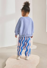 Hellblaues Sweatshirt mit Ballonärmel, kombiniert mit pink- und blau gestreiften Hosen mit Wellenmuster. Weiße Sneaker runden das Outfit ab.