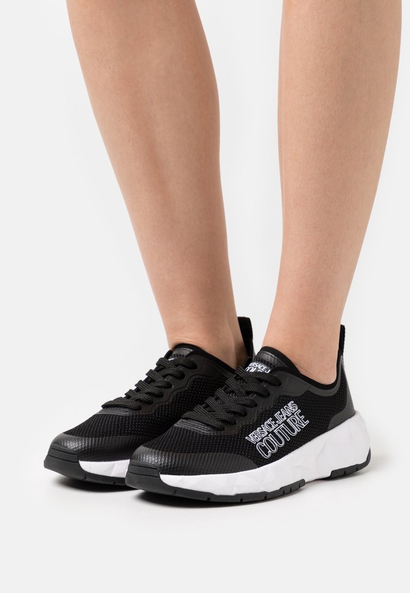 Svarta mesh-sneakers med vit gummisula och snören, med 'Versace Jeans Couture'-logotyp på sidan för en sportig stil.