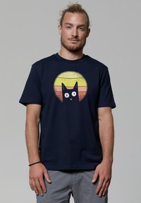 T-shirt en coton bleu marine avec un graphisme d'un chat noir sur un motif de cercles jaunes et orange usés. Manches courtes, col rond.