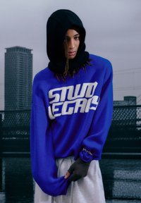 Blauwe trui met witte "STUDIO" tekst, zwarte capuchon en geribbelde manchet; gecombineerd met grijze joggingbroek en vingerloze handschoenen.