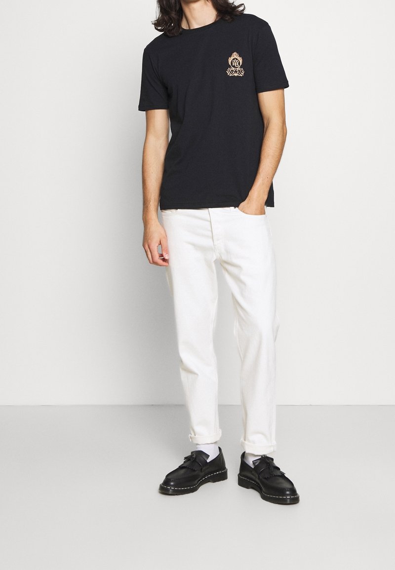 Homme portant un t-shirt noir avec un petit emblème sur la poitrine, un pantalon blanc retroussé, des chaussettes blanches et des chaussures en cuir noires, debout avec une main dans la poche.