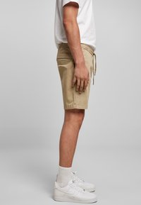 Personne portant un short beige, des chaussettes blanches, des baskets blanches et un t-shirt blanc, debout de profil sur un fond gris neutre.
