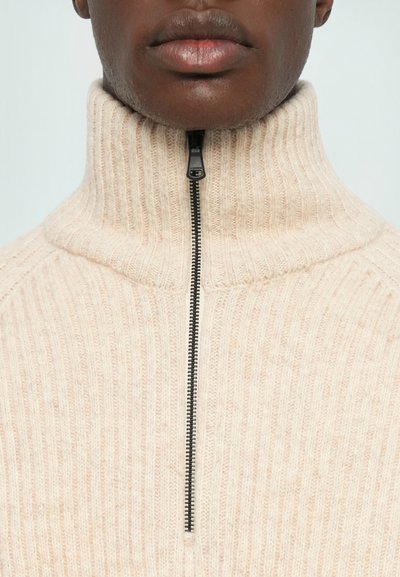 Beige ribbad stickad tröja med hög krage och delvis dragkedja. Mjuk textur med en framträdande dragkedja och ett enhetligt vertikalt mönster.