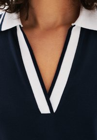 Granatowa koszulka polo z białym kołnierzykiem w paski. Posiada dekolt w serek i teksturowany materiał. Szczegóły obejmują kontrastowe obszycia wzdłuż brzegów.