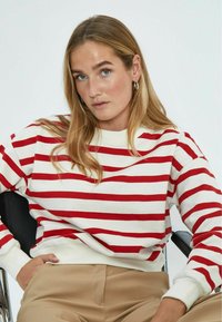 Felpa a righe rosse e bianche con polsini e scollo a coste; abbinata a pantaloni beige; modello seduto su una sedia nera.