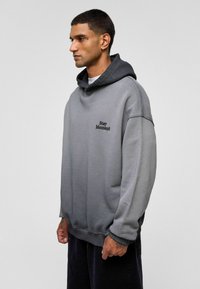 Grijze oversized hoodie met een gestructureerde afwerking, voorzien van een contrasterende zwarte capuchon en geborduurde "Stay Moment" detail op de borst.