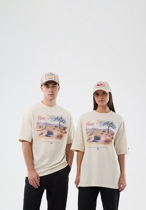 Vyras ir moteris stovi šalia vienas kito, dėvėdami smėlio spalvos grafinius marškinėlius ir atitinkančias kepures su dykumos peizažu ir užrašu „New Era“.