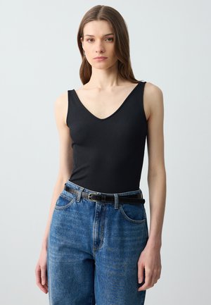 Jeune femme portant un haut noir sans manches à col en V rentré dans un jean bleu taille haute avec une ceinture noire, debout devant un fond uni clair.