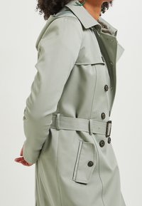 Světle zelený trenčkot s páskem v pase, dvouřadovým zapínáním, ramenními epauletami a bočními kapsami. Hladká, strukturovaná tkanina.