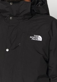 Veste noire North Face avec col haut, poche zippée sur la poitrine et logo brodé blanc sur le côté gauche.