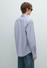 Camicia a maniche lunghe a righe azzurro chiaro con colletto abbottonato, caratterizzata da una vestibilità ampia e polsini con bottoni, indossata sopra pantaloni in denim scuro.