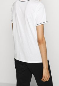 Persona vestida con una camisa de manga corta blanca con ribetes negros en las mangas y pantalones negros, vista desde atrás contra un fondo sencillo.