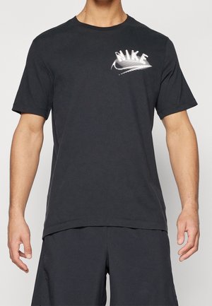 Homme portant un t-shirt Nike noir avec un logo Nike blanc déformé sur la poitrine gauche, assorti à un short noir, sur un fond uni.