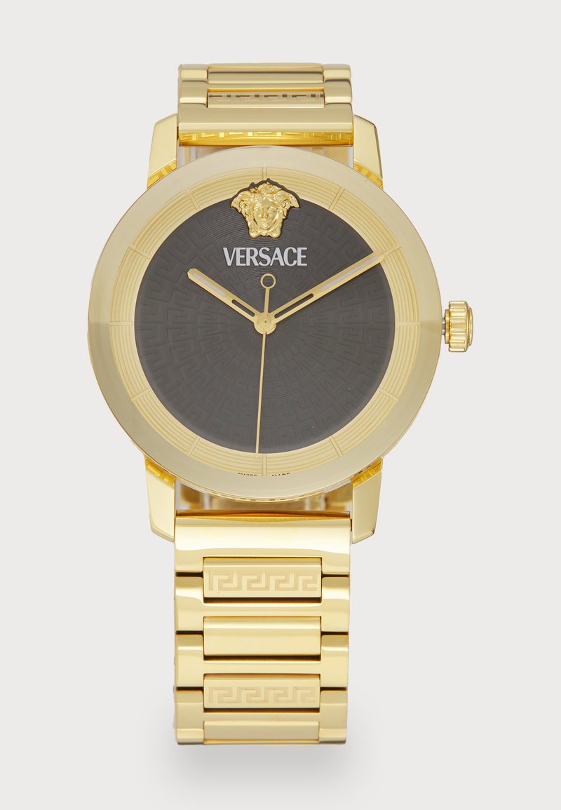 Kuldne analoogkell musta mustriga dials, millel on lõvi pea logo ja graveeritud detailid kuldsel käevõrul. Ümmargune kera disain.