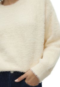 Un pull fluffy beige clair avec une texture douce, un col rond et des manches longues, associé à un jean en denim foncé.