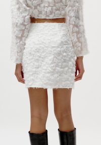 Mini-jupe blanche à texture avec un design en tissu effiloché. Assortie à un haut transparent à manches longues avec des détails effilochés correspondants. Bottes noires jusque aux genoux.