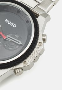 Hõbemetallist roostevabast terasest kell, millel on must tekstureeritud ääre, must kellanägu, punased aktsendid ja reljeefne "HUGO" logo küljes.