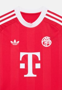 Camiseta deportiva roja con ribetes blancos, que presenta tres rayas blancas en los hombros, un gran logo blanco y un logo blanco más pequeño en el pecho.
