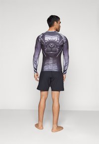 Zwarte lange mouwen compressie shirt met grijze leeuw graphic; gecombineerd met zwarte shorts, gedragen door een persoon, blootsvoets, bekeken van achteren.