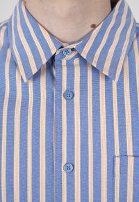 Gros plan d'une chemise bleue avec des rayures verticales jaunes et blanches, boutonnée au col, montrant le cou d'une personne avec un grain de beauté visible.
