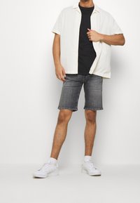 Camicia a maniche corte color crema con bottoni sopra una maglietta nera, abbinata a pantaloni shorts in denim scuro e sneakers bianche. Palette di colori neutri, design casual.