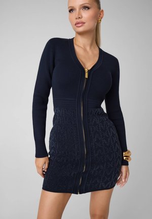 Femme portant une robe moulante bleu marine à manches longues avec une fermeture éclair dorée sur le devant et un motif texturé, des boucles d'oreilles créoles dorées et un bracelet épais doré.