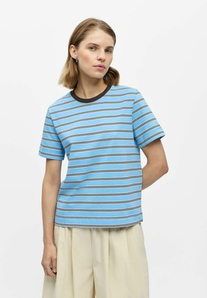 Jeune femme portant un t-shirt à manches courtes bleu et marron rayé et un pantalon large crème, posant devant un fond blanc uni.