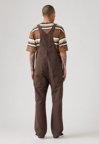 Man met tatoeages in een bruine overall en een gestreepte bruine, witte en zwarte korte mouw shirt, staand met zijn rug naar de camera tegen een effen achtergrond.