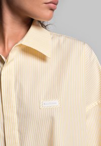 Gestreept geel shirt met een wit label; heeft een klassieke kraag, een gladde textuur en een losse pasvorm. De strepen zijn gelijkmatig verdeeld.
