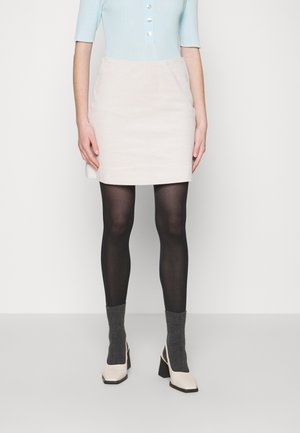 Woman wearing light blue top, cream-colored mini skirt, black tights, gray socks, and beige high-heeled shoes standing against a plain background.