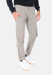 Pantaloni cargo grigio chiaro con molteplici tasche, dettagli cuciti rossi, un rinforzo imbottito sulle ginocchia e orli regolabili. Indossati con sneakers bianche.