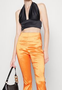 Svart satin halterneck crop top matchat med glänsande oranga utsvängda byxor. Outfiten har en slät textur och en svart handväska med kedjeaccent.