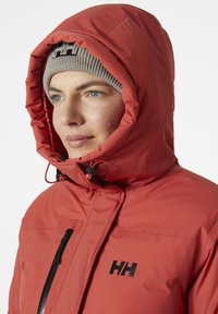 Chaqueta aislante roja con un alto cuello, capucha ajustable y bolsillo negro con cremallera. Presenta una textura suave y un logotipo prominente en el pecho.