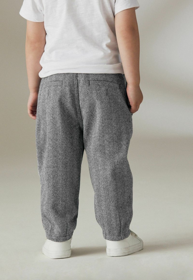 Enfant debout, de dos, portant un t-shirt blanc, un pantalon gris à motif chevrons et des baskets blanches sur un fond uni.