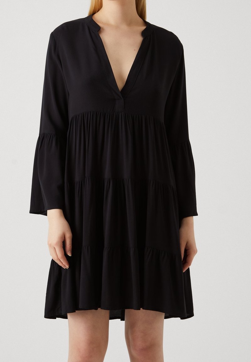 Robe noire à manches longues avec jupe à volants et col en V portée par une personne aux bras et jambes visibles sur un fond clair uni.