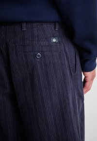 Navy pinstriped trousers with a button closure, side pockets, and a back pocket featuring a logo label. Textured fabric and tailored fit.