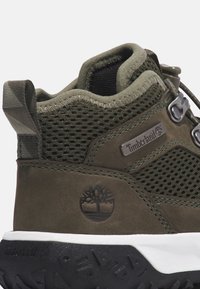 Ciemnozielone buty trekkingowe z siateczkowymi panelami, materiałem z zamszu, białą podeszwą środkową, czarną gumową podeszwą zewnętrzną i metalowymi oczkami. Widoczne logo Timberland.